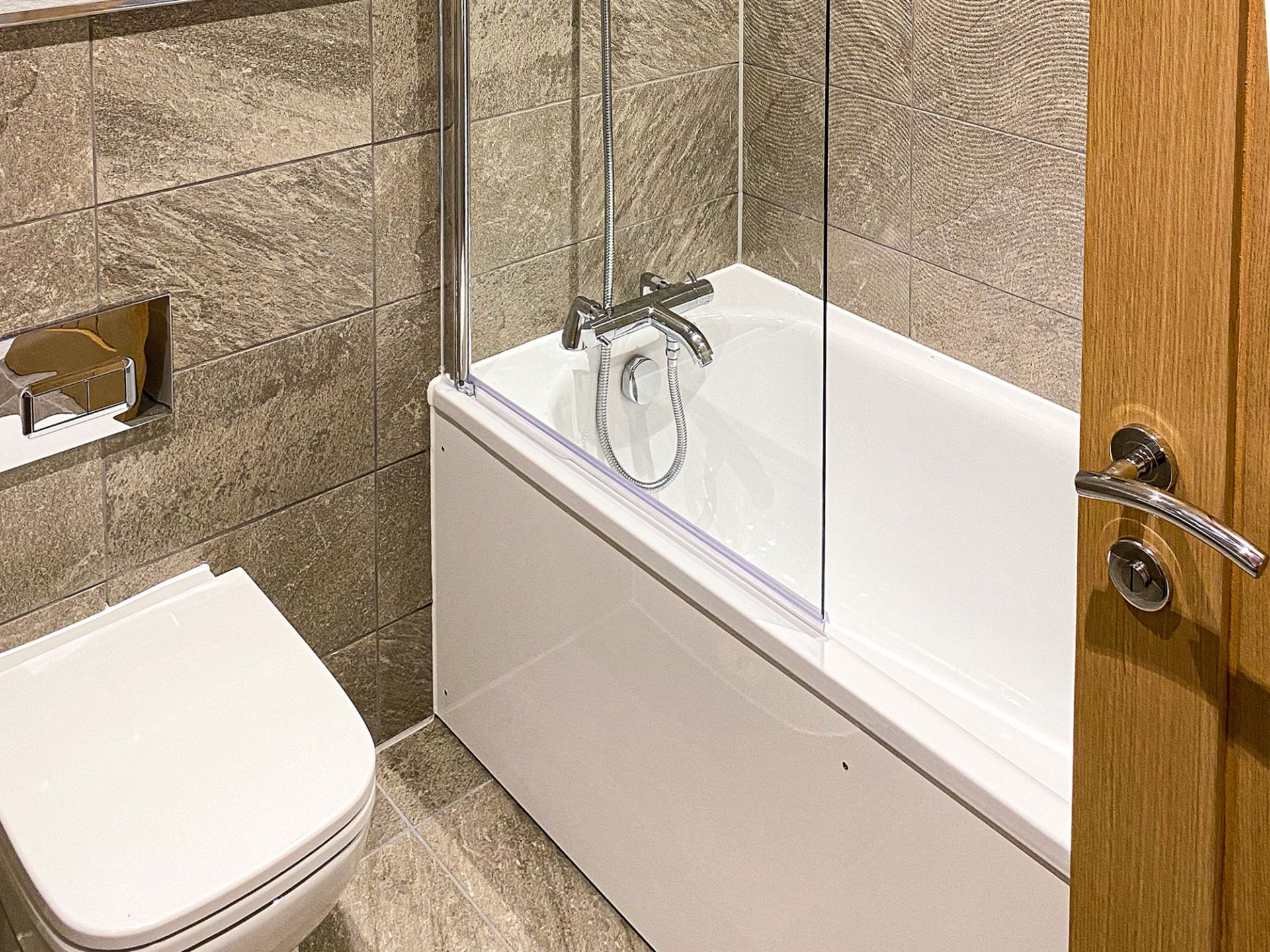 Finished bathroom in one of the townhouses. Slate grey patterned tile son walls and floor with white bath and wooden door.